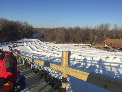 A snowy hill with tubing lanes, surrounded by trees, and a lodge in the background under a clear blue sky.