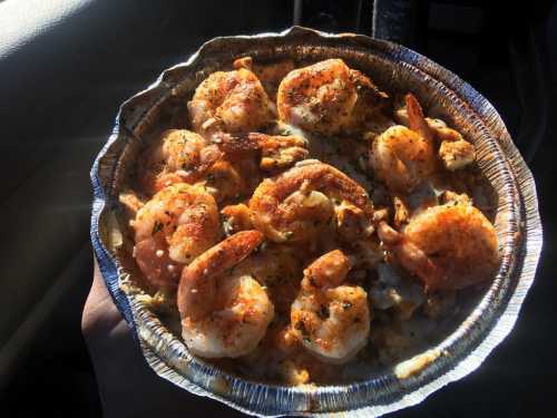 A hand holding a foil container filled with seasoned shrimp on a bed of rice, illuminated by sunlight.