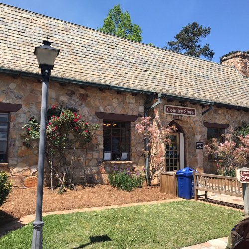A rustic stone building with a sign reading "Country Store," surrounded by greenery and flowers on a sunny day.