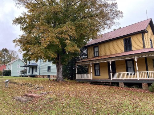 Two historic houses with porches, surrounded by autumn leaves and a large tree, set in a peaceful outdoor area.