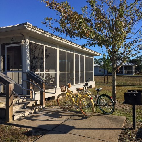 A screened porch with two bicycles parked outside, surrounded by grass and trees under a clear blue sky.
