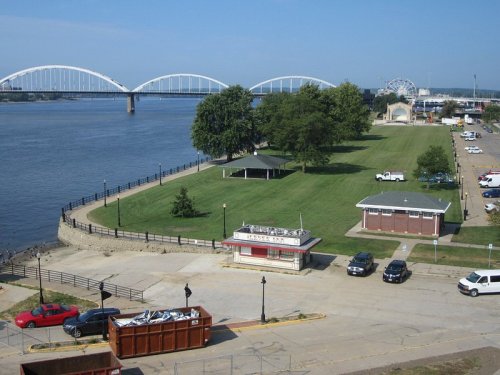 A riverfront view featuring a green park, a pavilion, and a bridge in the background under a clear blue sky.