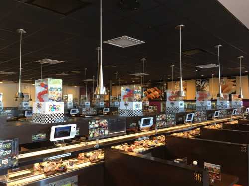 A spacious sushi restaurant with conveyor belts, digital menus, and colorful food displays under modern lighting.