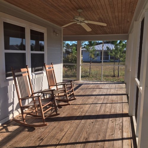 A porch with two wooden rocking chairs, a ceiling fan, and a view of a grassy area outside.