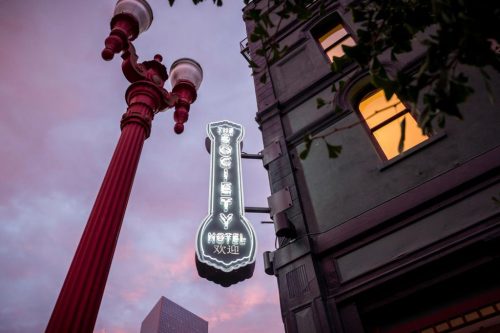 Neon sign for Society Hotel illuminated against a pink sky, with a vintage street lamp nearby.