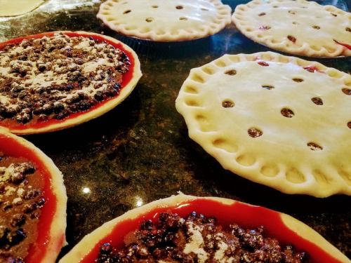 A countertop with unbaked pies, some filled with fruit and others with a plain crust, all with decorative edges.