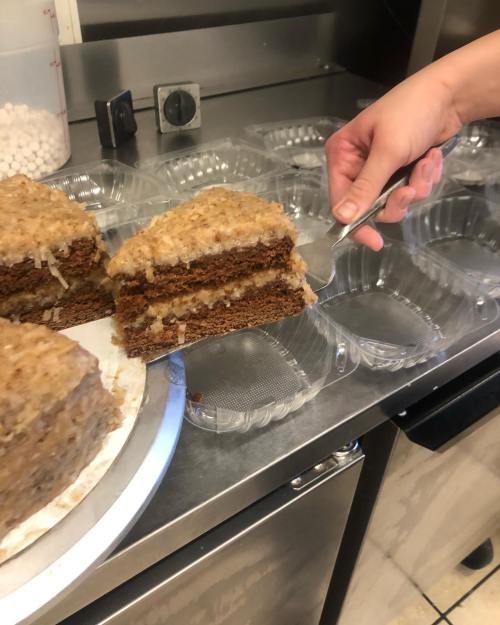 A hand serves a slice of layered cake from a tray, with additional cake pieces in plastic containers nearby.