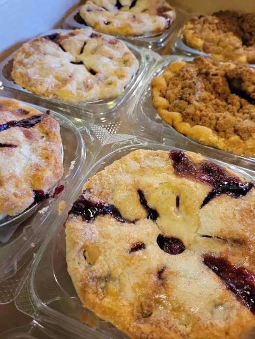 A variety of freshly baked pies in clear containers, featuring fruit fillings and crumb toppings.