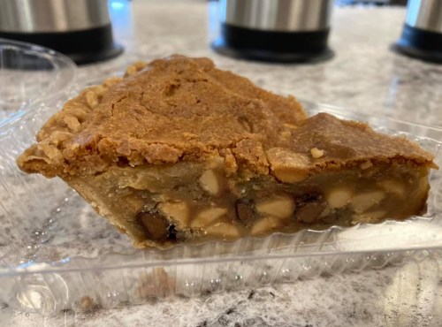 A slice of cookie pie with nuts, resting in a clear plastic container on a marble countertop.