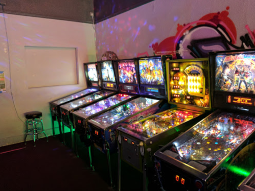 A row of colorful pinball machines illuminated by disco lights in a vibrant arcade setting.