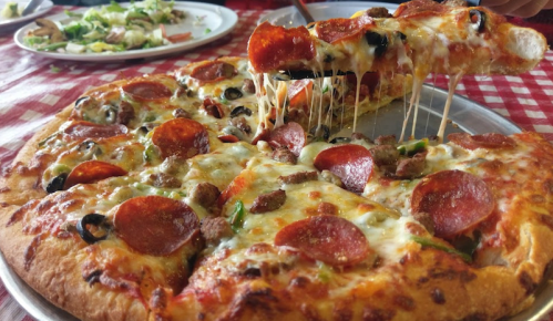 A close-up of a cheesy pepperoni pizza with toppings, being pulled apart, on a checkered tablecloth.