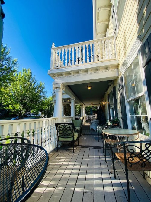 A sunny porch with white railings, green chairs, and a table, surrounded by trees and a clear blue sky.