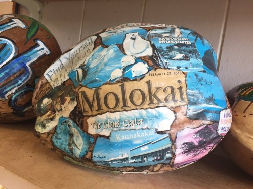 A decorated stone featuring images and text about Molokai, including landmarks and a date, displayed on a shelf.
