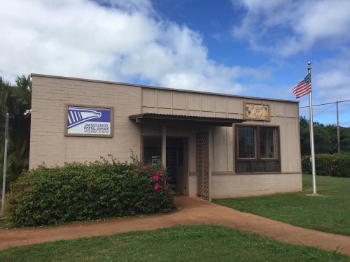 A small post office building with a sign, surrounded by greenery and an American flag flying nearby.
