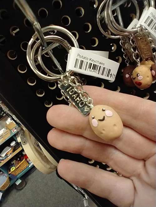 A hand holding a cute potato keychain with a "Idaho" tag, displayed on a pegboard with other keychains.