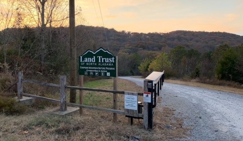 Sign for the Land Trust of North Alabama at a nature preserve, with a gravel road and wooded hills in the background.