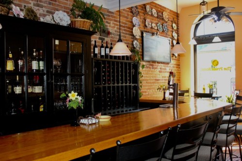 A cozy bar with a wooden counter, shelves of drinks, and brick walls adorned with decorative plates.