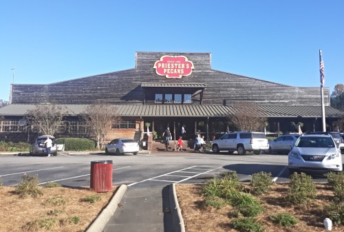 A rustic building with a sign reading "Priester's Pecans," surrounded by parked cars and trees under a clear blue sky.