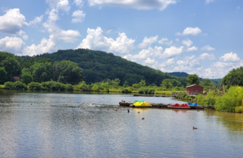 A serene lake scene with colorful paddle boats, lush greenery, and a hill under a partly cloudy sky. Ducks swim nearby.