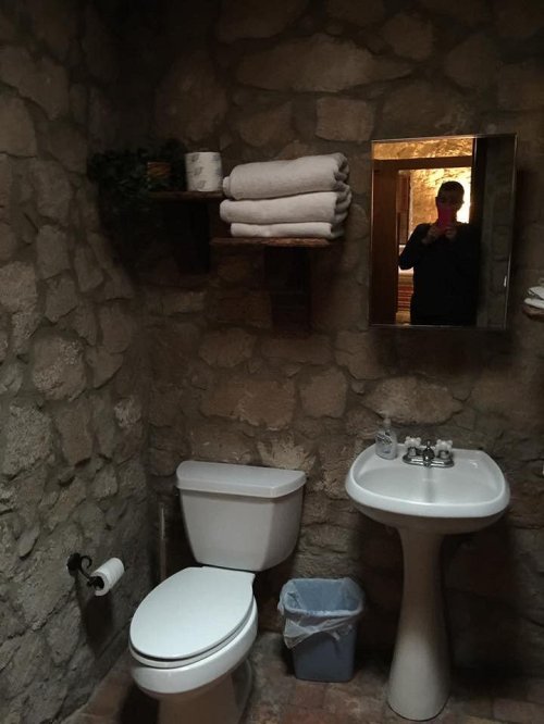 A rustic bathroom with stone walls, featuring a toilet, sink, towels on a shelf, and a mirror reflecting a person.