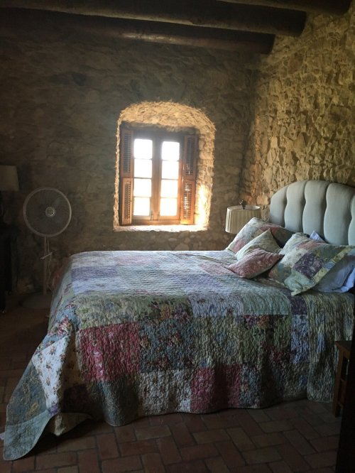 Cozy bedroom with a stone wall, large window, and a colorful quilted bedspread. A fan is visible in the corner.