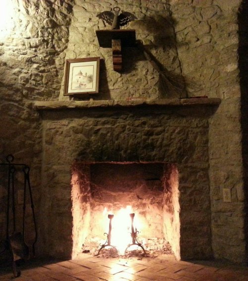 A cozy stone fireplace with a warm fire, framed by a rustic wall and a shelf holding a picture and a broom.