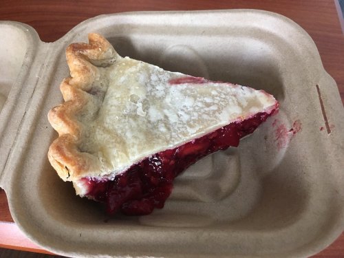 A slice of cherry pie in a brown, eco-friendly takeout container. The crust is golden and flaky, with a glossy filling.