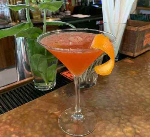 A vibrant cocktail in a martini glass, garnished with an orange twist, sits on a bar counter with greenery in the background.