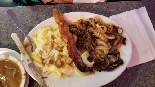 A plate of meat with onions, crispy bacon, and mashed potatoes with gravy on a textured table surface.