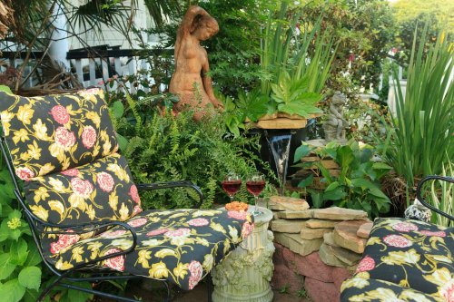 A cozy garden scene with floral chairs, a statue, and two glasses of red wine amidst lush greenery.