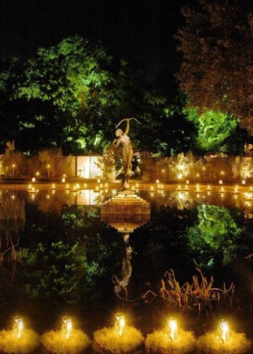 A serene night scene featuring a statue by a pond, surrounded by glowing candles and lush greenery.
