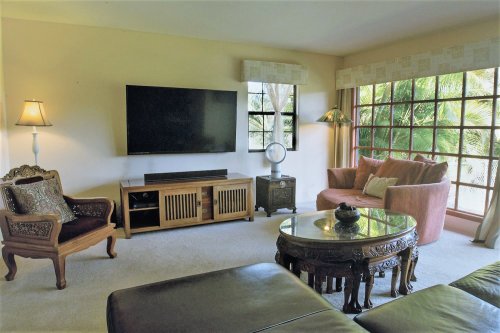 A cozy living room with a large TV, a brown sofa, an ornate chair, and a round coffee table by a window with greenery outside.
