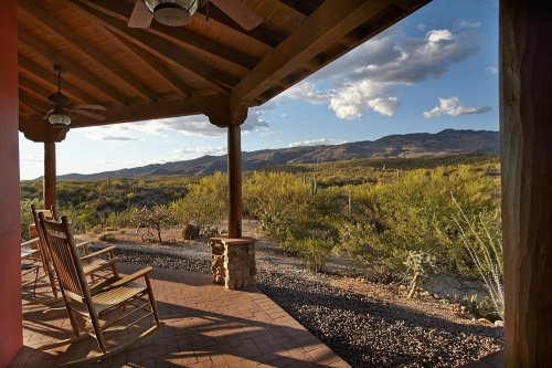 A porch with rocking chairs overlooks a desert landscape and mountains under a blue sky with scattered clouds.