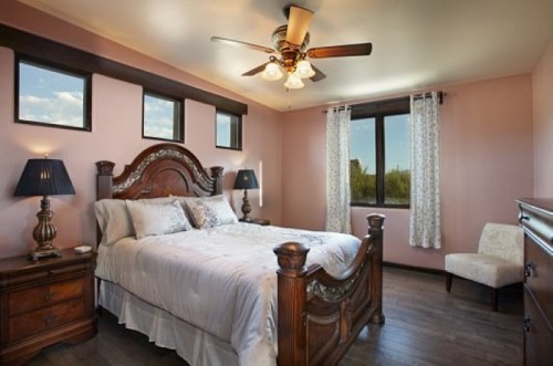 Cozy bedroom with a wooden bed, two lamps, and large windows, featuring light pink walls and wooden flooring.