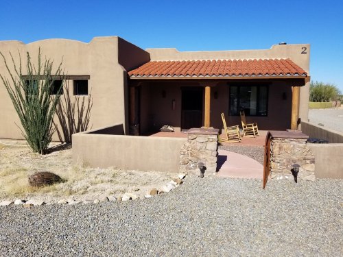 A southwestern-style house with a red tile roof, stone accents, and rocking chairs on the porch, surrounded by desert landscape.