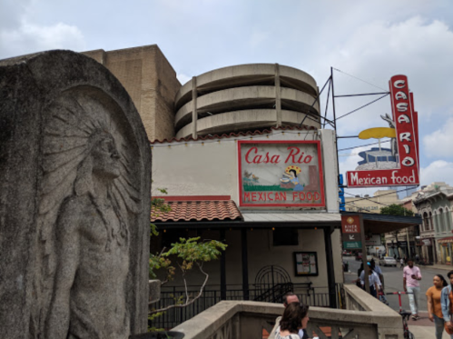 A stone statue stands in front of Casa Rio, a Mexican restaurant with a colorful sign, in a bustling urban area.