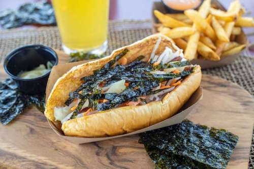 A hot dog topped with vegetables and seaweed, served with fries and a drink on a wooden platter.