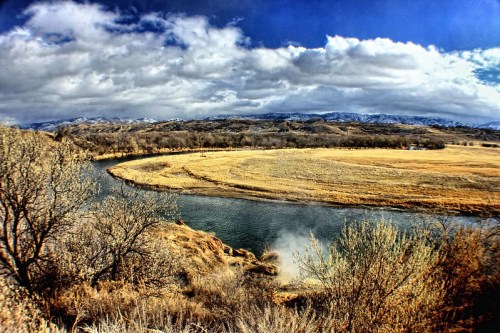 A winding river flows through a landscape of dry grass and trees, with mountains and clouds in the background.