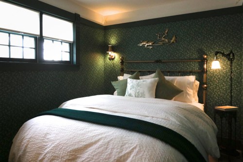 Cozy bedroom with a patterned green wallpaper, a bed with decorative pillows, and soft lighting.