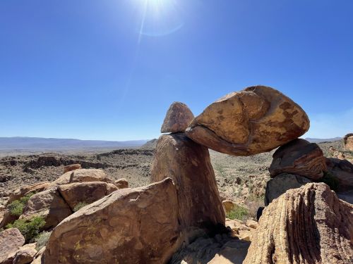 A large rock balanced on top of another rock, set against a clear blue sky and desert landscape.