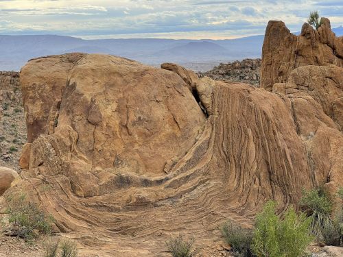 A large, textured rock formation with layered patterns, set against a cloudy sky and distant mountains.