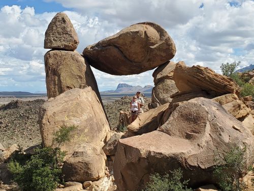 A person stands near large, balanced rock formations against a backdrop of mountains and a cloudy sky.
