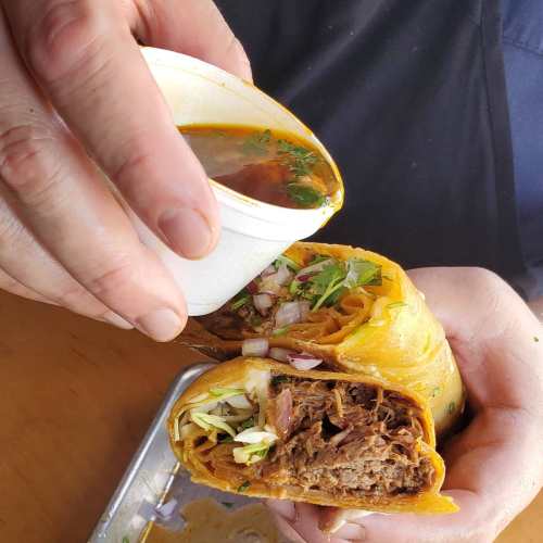 A hand holds a small cup of sauce above two rolled tacos filled with meat and garnished with onions and cilantro.