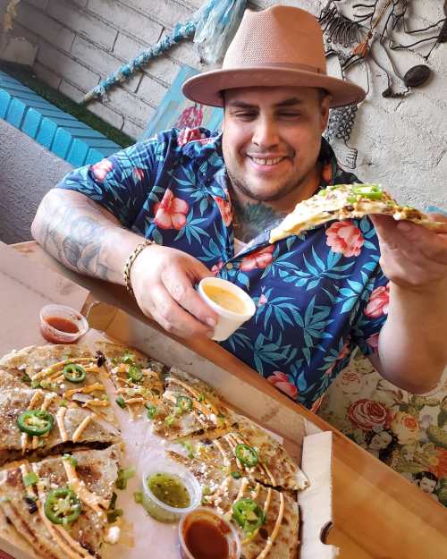A smiling man in a floral shirt holds a slice of pizza over a large pizza with sauces on the side.