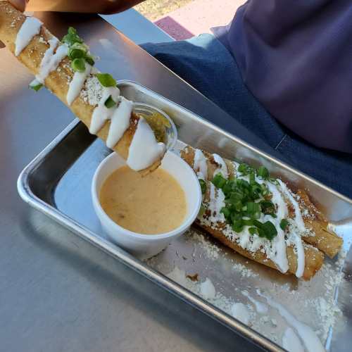 A plate of crispy taquitos topped with sour cream and green onions, served with a side of dipping sauce.