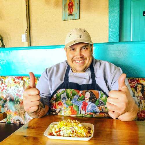 A smiling chef in a colorful apron gives a thumbs up while sitting at a table with a plate of food.