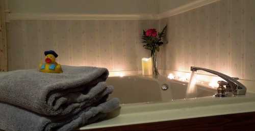 A cozy bathroom scene with a rubber duck, stacked towels, a candle, and a vase of roses by a bathtub.