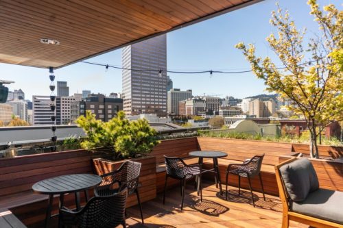 A rooftop terrace with wooden seating, overlooking a city skyline and tall buildings under a clear blue sky.