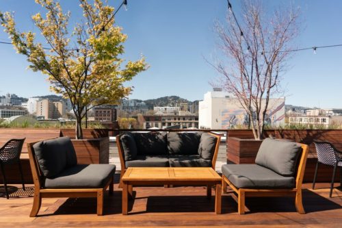 A rooftop patio with wooden furniture, including a couch and table, surrounded by trees and a city skyline in the background.
