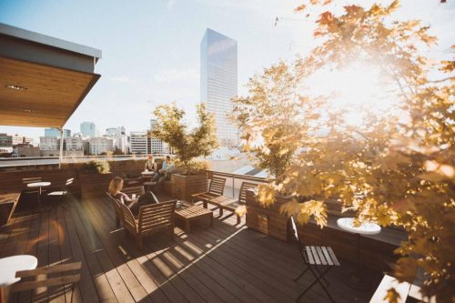 A sunlit rooftop terrace with wooden seating, plants, and a city skyline in the background.